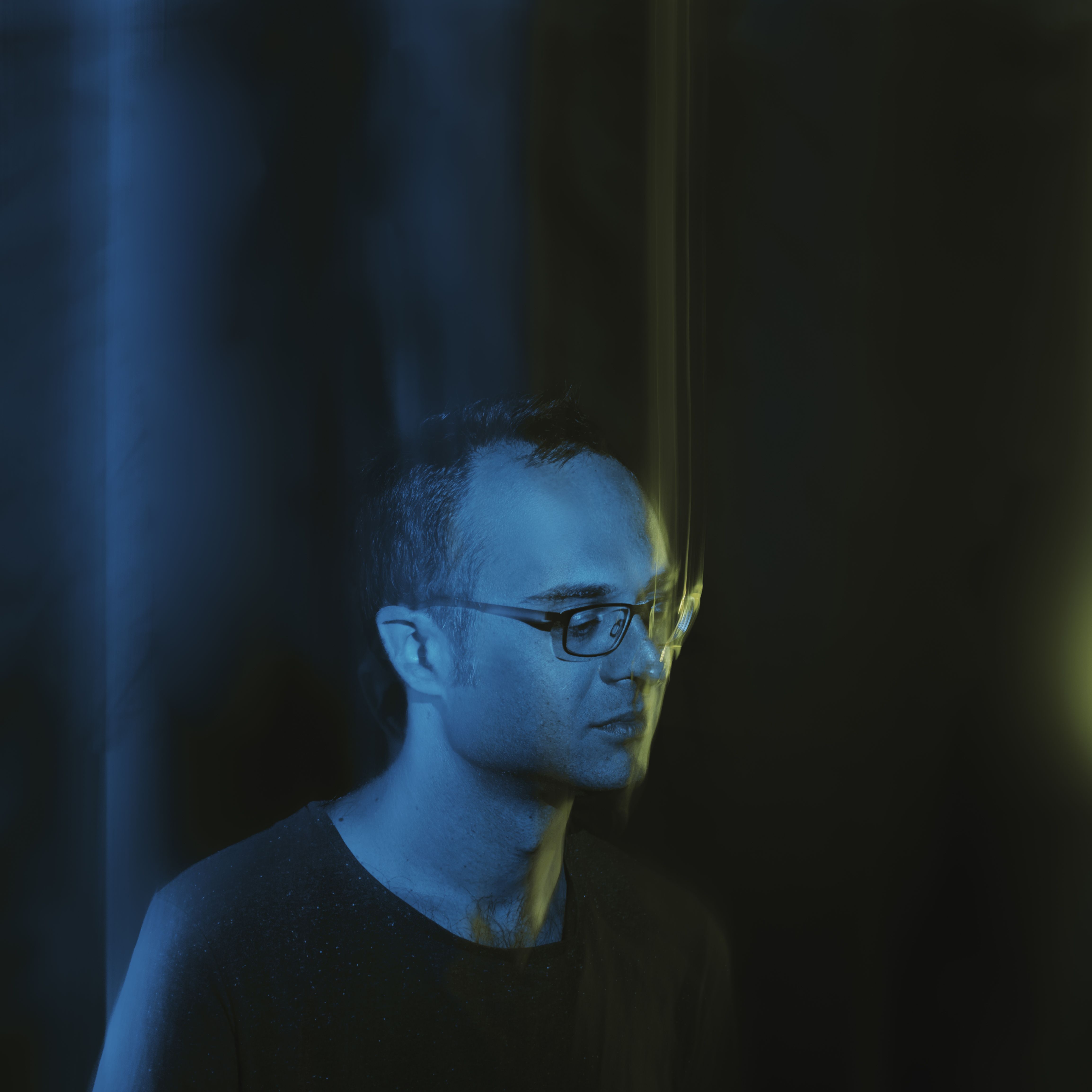 black background with blue swish light with man in black shirt wearing glasses sitting down