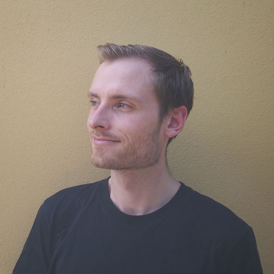 Photo of man standing against stone wall with short brown hair and black shirt. staring away from camera into distance